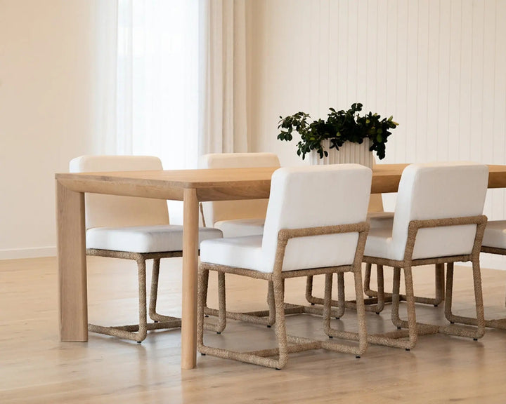 A light oak dining table with four matching chairs featuring white upholstered seats and natural woven rope armrests, set in a bright, modern dining room with a potted plant on the table. Aire Dining Table | Rustic Charm Interiors 