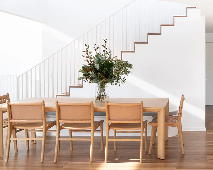 A modern dining room scene with a light wood dining table and four matching woven-back chairs, set in a bright white interior with a staircase and hardwood floor. Aire Dining Table | Rustic Charm Interiors 