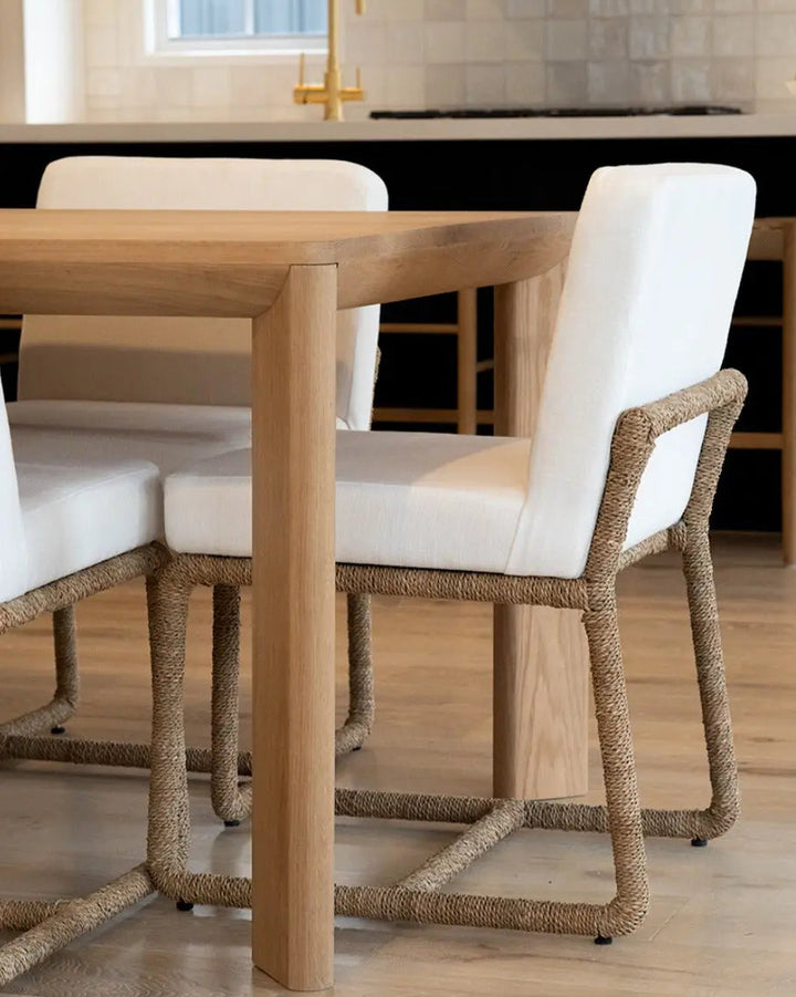 A dining chair with a white upholstered seat and back and a natural woven rope frame, shown beside a light wood dining table in a modern kitchen setting.