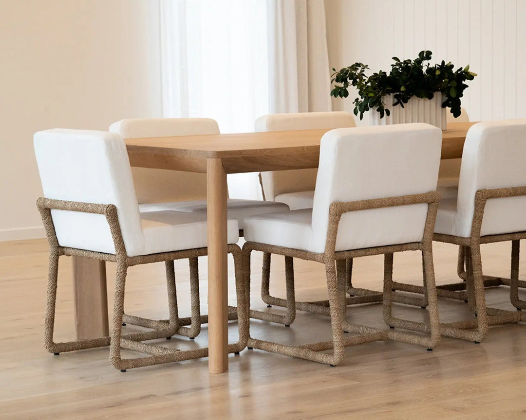 A dining set featuring a light wood table and six matching chairs with white upholstered seats and rope-wrapped wooden frames, set in a bright dining room with a potted plant in the background. Aire Dining Table | Rustic Charm Interiors 