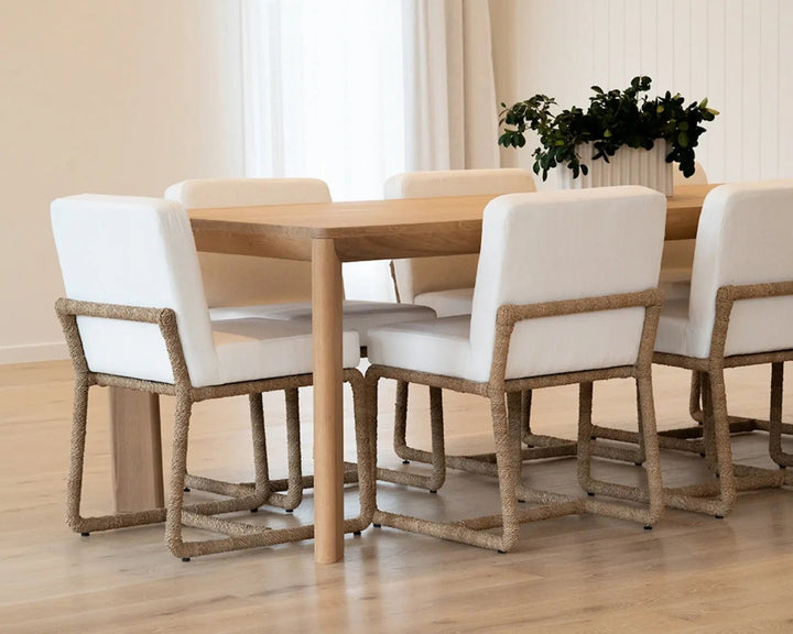 A dining set featuring a light wood table and six matching chairs with white upholstered seats and rope-wrapped wooden frames, set in a bright dining room with a potted plant in the background. Aire Dining Table | Rustic Charm Interiors 