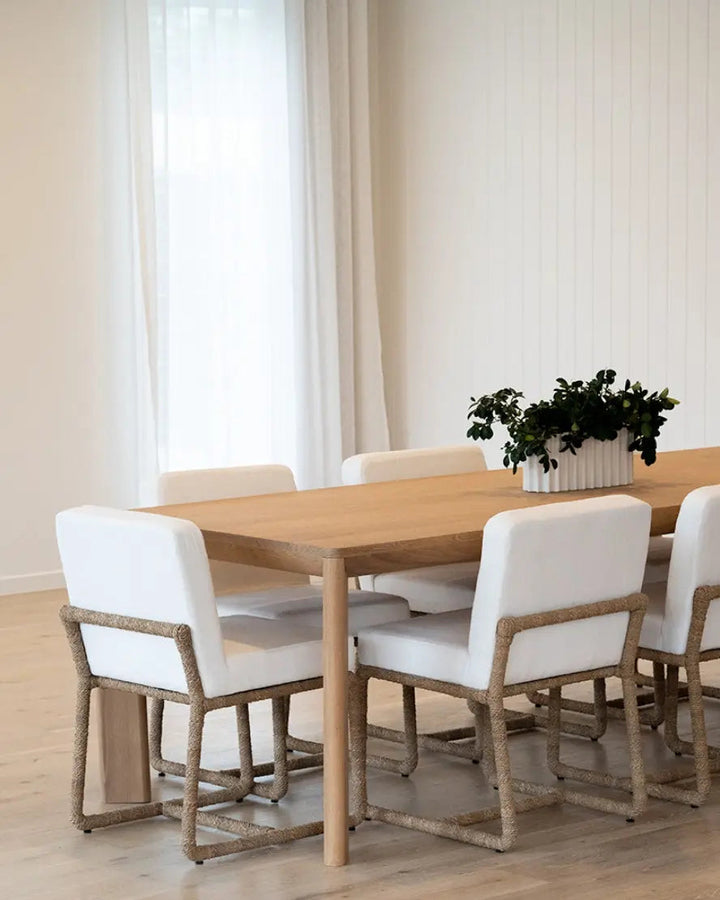 A modern dining table set in a bright, minimalist dining room. The set includes a rectangular light oak wood table with a natural finish and six matching dining chairs featuring white upholstered seats and natural woven rope frames.