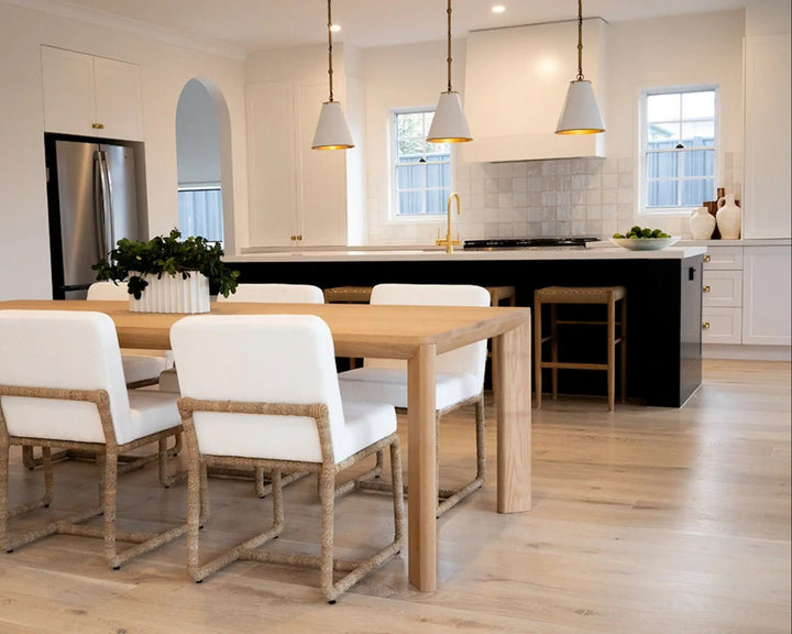 A modern dining room scene featuring a light wood dining table with six white upholstered chairs that have rope-wrapped frames, set in a bright, contemporary kitchen with light wood floors and white cabinetry. Aire Dining Table | Rustic Charm Interiors 