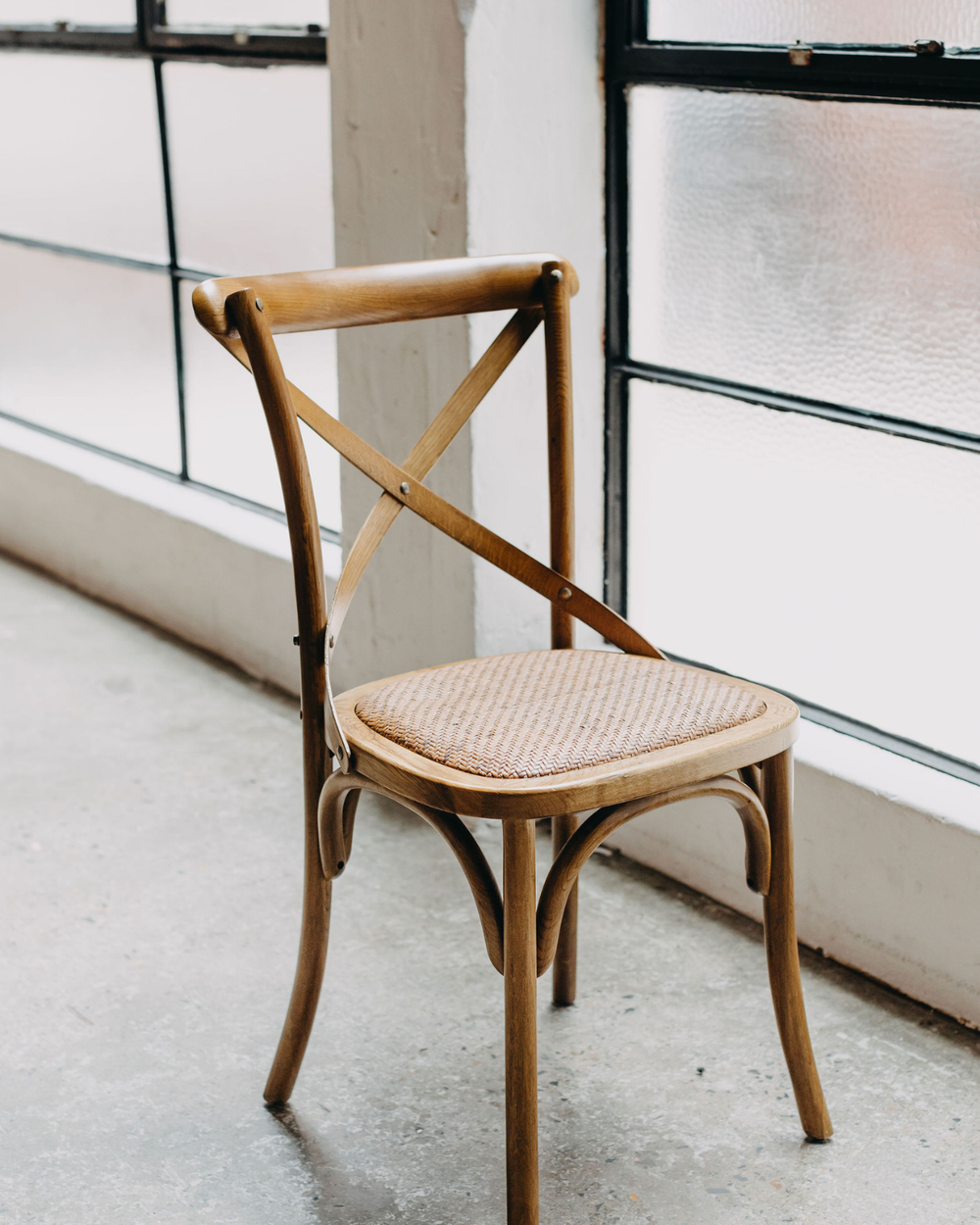 A wooden dining chair with a woven cane seat, positioned in front of large industrial windows with frosted glass.