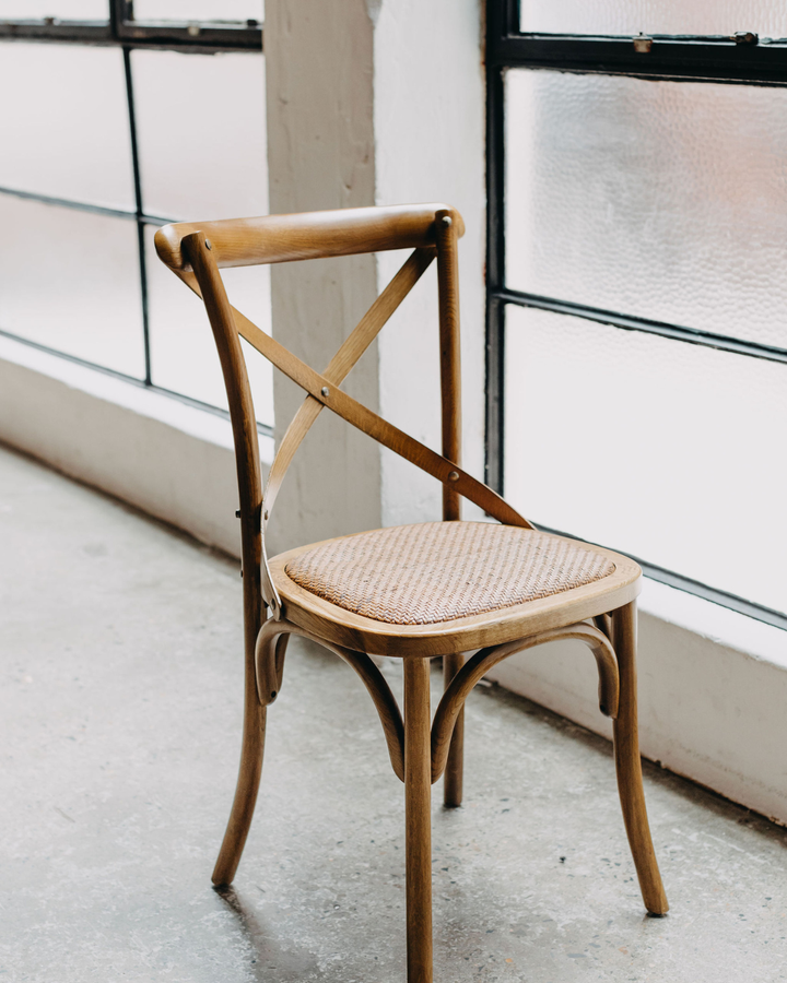 A wooden dining chair with a woven cane seat, positioned in front of large industrial windows with frosted glass.