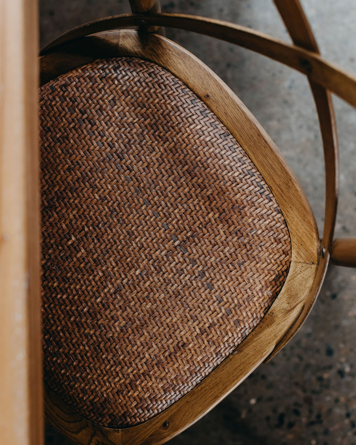 Close-up of a wooden chair seat with a woven brown rattan or wicker pattern, framed by light natural wood, on a concrete floor.