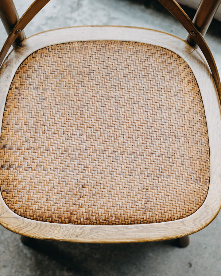 Close-up of a woven rattan or cane chair seat with a light natural wood frame, showing the detailed texture of the seat and part of the wooden backrest.