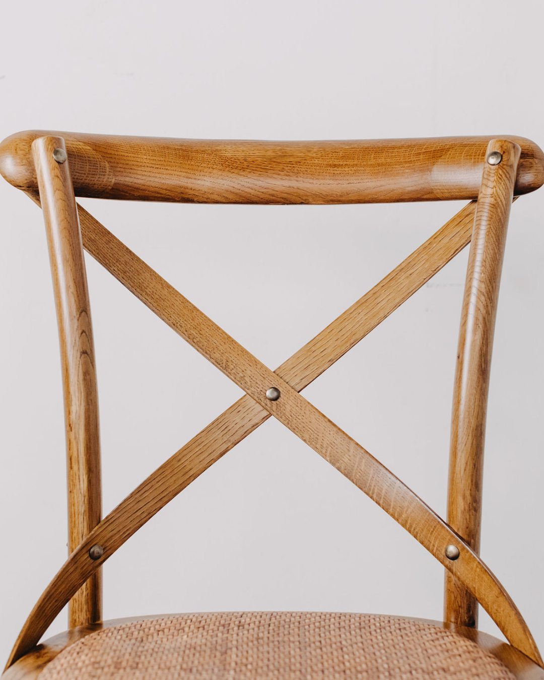 Close-up of a wooden dining chair back with a curved top rail, crossed X-shaped support slats, and a woven seat, shown against a plain light gray background.