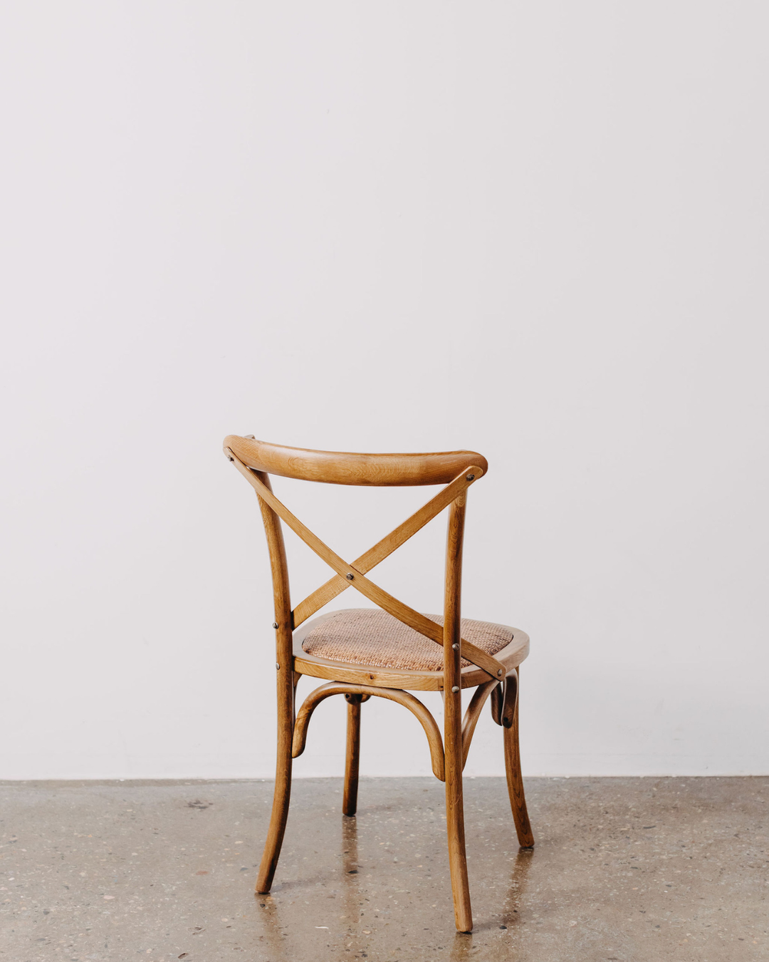 A light brown wooden dining chair with a woven cane seat, shown from the back against a minimalist white wall and concrete floor.