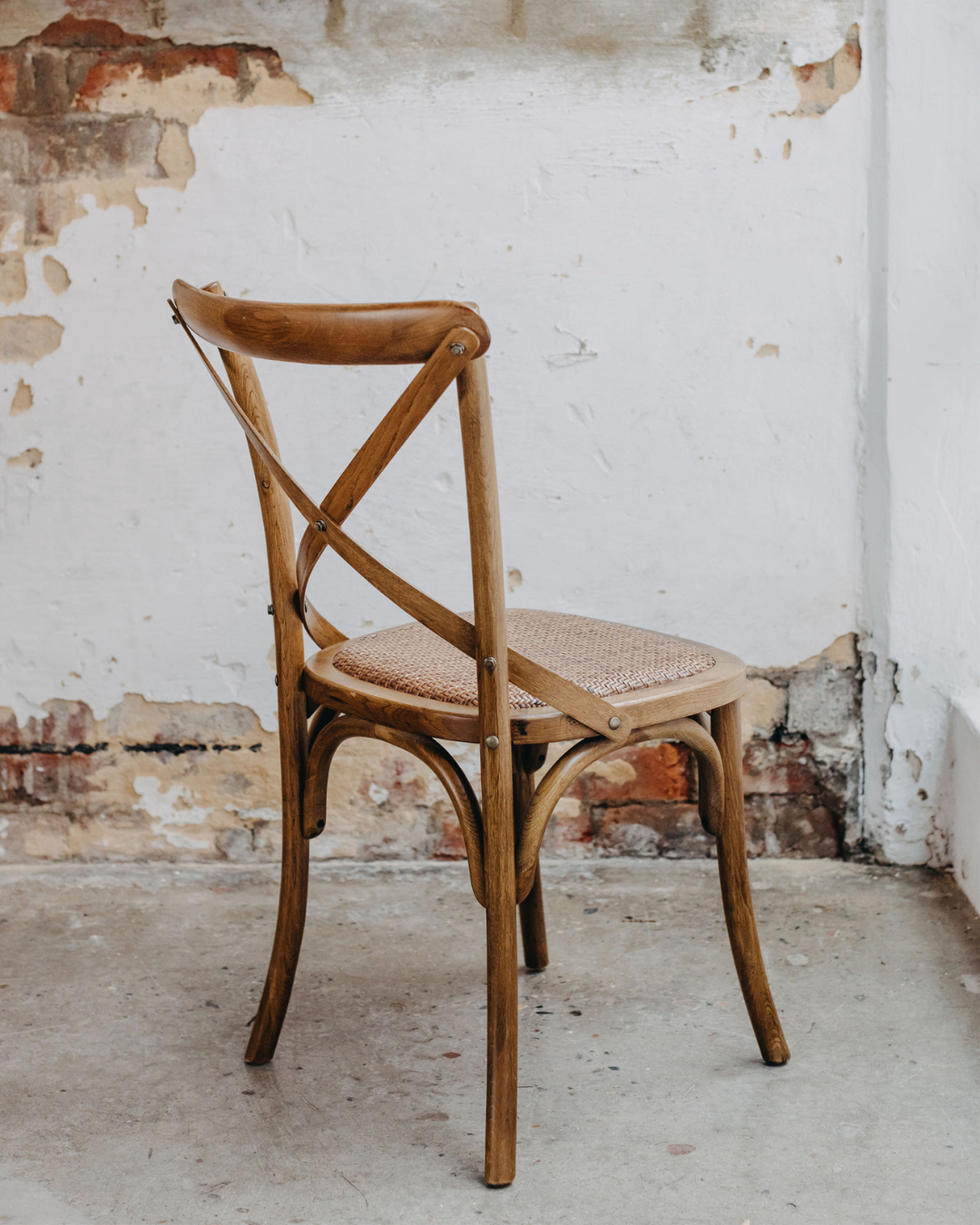 A classic wooden dining chair with a woven cane seat, positioned against a rustic white plaster wall with exposed brick detailing.