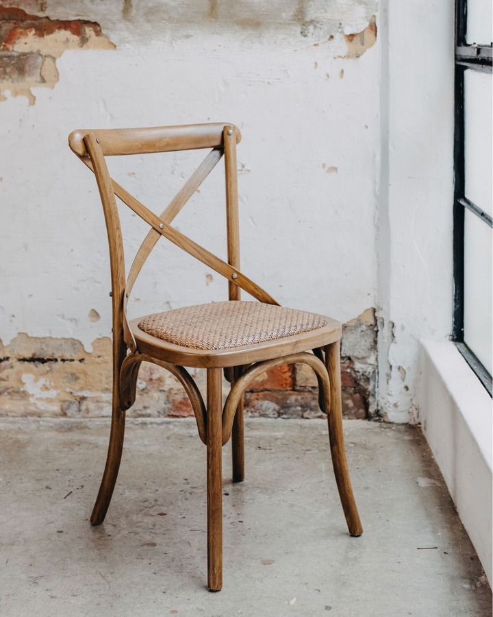 A rustic wooden dining chair with a woven cane seat, positioned in front of a distressed white wall with exposed brick and a concrete floor.