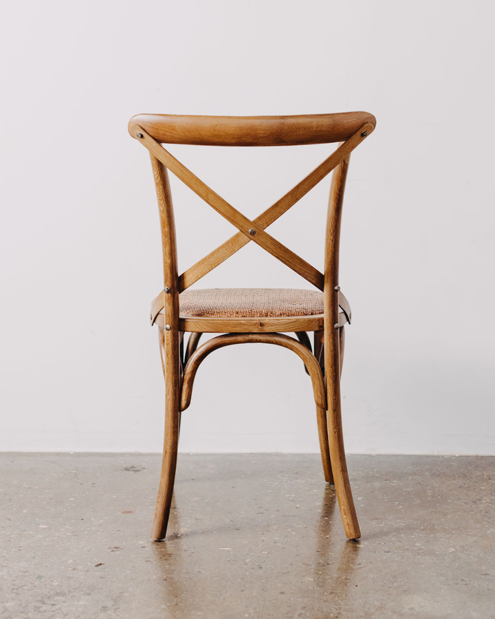 A vintage wooden dining chair with a curved backrest, crossed X-back design, and a woven seat cushion, shown against a plain light gray wall and concrete floor.