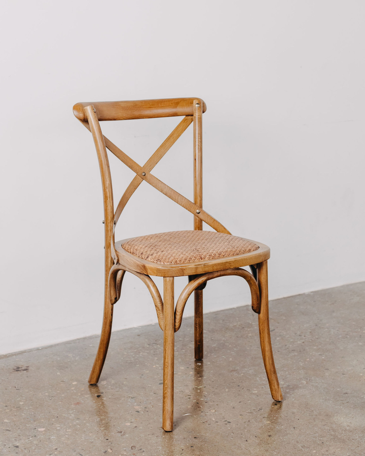 A vintage-style wooden dining chair with a curved bentwood frame, crossed backrest slats, and a woven cane seat, shown on a concrete floor against a plain light gray wall.