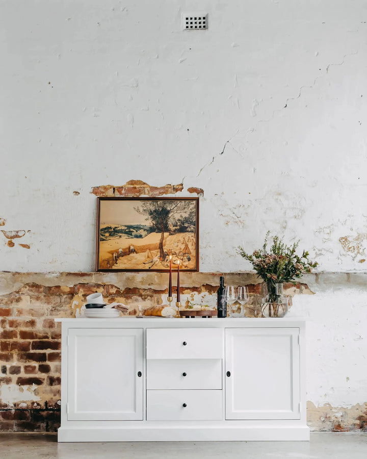A white wooden sideboard or credenza with three drawers and two cabinet doors, styled against a distressed white and exposed brick wall. A framed landscape painting and a vase of flowers sit on top, with a bottle and glasses in front.