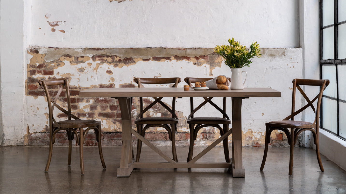 A rustic dining room scene featuring a light wood rectangular table with a trestle base, surrounded by four dark wood chairs. A white ceramic vase with yellow flowers sits on the table.